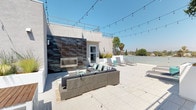 This rooftop terrace offers a well-appointed outdoor living experience designed for both relaxation and entertaining. The expansive deck is finished with large-format light grey porcelain tiles, providing a clean, modern aesthetic throughout the space.

A striking accent wall clad in textured, oxidized metal panels serves as the focal point of the lounge area, housing a sleek built-in linear fireplace. In front of the fireplace, a generously sized wicker sectional sofa with charcoal cushions and teal and striped accent pillows creates an inviting gathering space, complemented by a patterned area rug and a coffee table.

On the opposite end of the terrace, two contemporary adjustable sun loungers are positioned for sunbathing, accompanied by a teal accent table and decorative accessories. Raised planters featuring ornamental grasses add a natural, softening element to the otherwise modern design.

String lights are strung overhead across the terrace, enhancing the ambiance for evening use. Glass panel railings along the perimeter maintain unobstructed panoramic views of the surrounding neighborhood, including mature trees and the open sky. The overall atmosphere is bright, airy, and sophisticated.