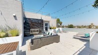This rooftop terrace offers a well-appointed outdoor living experience designed for both relaxation and entertaining. The expansive deck is finished with large-format light grey porcelain tiles, providing a clean, modern aesthetic throughout the space.

A striking accent wall clad in textured, oxidized metal panels serves as the focal point of the lounge area, housing a sleek built-in linear fireplace. In front of the fireplace, a generously sized wicker sectional sofa with charcoal cushions and teal and striped accent pillows creates an inviting gathering space, complemented by a patterned area rug and a coffee table.

On the opposite end of the terrace, two contemporary adjustable sun loungers are positioned for sunbathing, accompanied by a teal accent table and decorative accessories. Raised planters featuring ornamental grasses add a natural, softening element to the otherwise modern design.

String lights are strung overhead across the terrace, enhancing the ambiance for evening use. Glass panel railings along the perimeter maintain unobstructed panoramic views of the surrounding neighborhood, including mature trees and the open sky. The overall atmosphere is bright, airy, and sophisticated.
