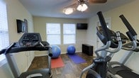 This dedicated fitness room offers a practical and comfortable space for a variety of workout routines. The room features durable wood-style flooring that provides both an attractive aesthetic and easy maintenance. Two windows along the back wall allow natural light to fill the space, creating a bright and energizing environment. A ceiling fan with an integrated light fixture helps maintain airflow and comfort during exercise sessions. A wall-mounted flat-screen television is positioned at eye level, ideal for entertainment or guided workout viewing. The room is equipped with a treadmill and an elliptical machine, catering to cardiovascular training needs. Exercise balls and yoga mats are also present, supporting stretching, balance, and core training. A low storage cube organizer provides convenient space for keeping fitness accessories organized and accessible. White chair rail molding runs along the walls, adding a subtle architectural detail. A door on the right side of the room suggests easy access to adjacent spaces. Overall, the room is thoughtfully arranged to maximize functionality while maintaining a clean, welcoming atmosphere suited for daily fitness use.