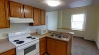 This single-floor space centers around a practical kitchen that serves as a key functional area. The kitchen is outfitted with warm-toned wood cabinetry both above and below the countertops, providing ample storage. A white electric coil range with an overhead ventilation hood anchors one side of the kitchen, while a double-basin stainless steel sink is positioned on an L-shaped counter configuration opposite. The countertops offer functional workspace between the appliances. A microwave sits conveniently on the counter, and under-cabinet storage is accessible below the sink area. The kitchen features a drop ceiling with a centrally mounted flush light fixture that provides overhead illumination. A window positioned near the sink area allows natural light to enter the space, brightening the kitchen and offering a view to the exterior. The flooring transitions from hard surface in the kitchen area to carpet in the adjacent room, visible at the edge of the frame. The walls are finished in a neutral light tone, contributing to a clean and straightforward atmosphere throughout the space.