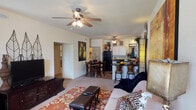 This single-floor residence offers an open-concept layout that seamlessly connects the living room, dining area, and kitchen into one cohesive living space. The living room is carpeted and comfortably sized, anchored by a ceiling fan with a light fixture that provides both airflow and ambient lighting. A patterned area rug and ample seating define the space, while a media console along one wall adds functional storage.

The dining area sits just off the living room, accommodating a full dining table and chairs, and transitions naturally into the kitchen. The kitchen is equipped with full-sized appliances including a refrigerator and range, white cabinetry, and dark countertops. A breakfast bar with bar stool seating faces the living area, creating an informal dining and social zone that enhances the open feel of the layout.

Arched passageways connect the main living area to additional rooms, adding an architectural detail that gives the space a defined yet flowing character. The neutral wall tones throughout create a bright, clean backdrop. The overall ambiance is warm and livable, with a practical floor plan that makes efficient use of space while maintaining a comfortable and welcoming atmosphere suitable for everyday living.