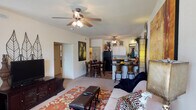 This single-floor residence offers an open-concept layout that seamlessly connects the living room, dining area, and kitchen into one cohesive living space. The living room is carpeted and comfortably sized, anchored by a ceiling fan with a light fixture that provides both airflow and ambient lighting. A patterned area rug and ample seating define the space, while a media console along one wall adds functional storage.

The dining area sits just off the living room, accommodating a full dining table and chairs, and transitions naturally into the kitchen. The kitchen is equipped with full-sized appliances including a refrigerator and range, white cabinetry, and dark countertops. A breakfast bar with bar stool seating faces the living area, creating an informal dining and social zone that enhances the open feel of the layout.

Arched passageways connect the main living area to additional rooms, adding an architectural detail that gives the space a defined yet flowing character. The neutral wall tones throughout create a bright, clean backdrop. The overall ambiance is warm and livable, with a practical floor plan that makes efficient use of space while maintaining a comfortable and welcoming atmosphere suitable for everyday living.