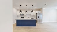 Located in Sugar Hill, Georgia, this well-appointed four-bedroom, three-bathroom home spans three floors, offering a generous and functional layout throughout. The entry-level features a concrete-floored garage space with windows that bring in natural light, providing ample utility. The main living floor is the heart of the home, anchored by an open-concept kitchen and living area. The kitchen is a standout, boasting white shaker cabinetry, a light granite countertop, a striking navy-blue island with seating space, stainless steel appliances, and three industrial-style pendant lights overhead — all set atop light hardwood flooring. The bathrooms are finished with wood-look tile flooring, white subway tile tub surrounds, granite-topped vanities, and brushed nickel fixtures. Bedrooms are carpeted and bright, each equipped with ceiling fans and windows offering neighborhood views. One bedroom features hardwood flooring, adding variety across the home. Closets include built-in wire shelving for practical storage. Multiple staircases connect the three floors seamlessly. The overall aesthetic is clean and modern, with a neutral palette of whites and warm beiges punctuated by bold design choices in the kitchen, creating a cohesive and inviting living environment.