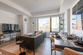 This thoughtfully designed single-floor urban residence showcases a refined contemporary aesthetic throughout. The open-concept living, dining, and kitchen area serves as the heart of the home, bathed in natural light through expansive floor-to-ceiling windows that frame striking city skyline views. Tray ceilings add architectural depth to the space, while wide-plank hardwood flooring flows seamlessly from room to room.

The kitchen is a standout feature, outfitted with two-tone cabinetry in light grey and dark wood, a veined marble-look backsplash, stainless steel appliances, and a generous waterfall-edge island with bar seating beneath a sculptural linear pendant light. A built-in wine rack and wet bar area add an elegant entertaining touch. The living and dining zones are anchored by a bold navy blue wall panel, creating a dramatic focal point.

The bedroom is a serene retreat with clean white walls, wood floors, and large windows overlooking the urban landscape. The space accommodates a full suite of furnishings with ease. The bathroom is finished to a high standard, featuring large-format porcelain tile flooring, a marble-topped vanity with undermount sink, backlit mirror, and a frameless glass walk-in shower with neutral tile surround. Every detail reflects a cohesive, upscale design sensibility.