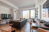 This thoughtfully designed single-floor urban residence showcases a refined contemporary aesthetic throughout. The open-concept living, dining, and kitchen area serves as the heart of the home, bathed in natural light through expansive floor-to-ceiling windows that frame striking city skyline views. Tray ceilings add architectural depth to the space, while wide-plank hardwood flooring flows seamlessly from room to room.

The kitchen is a standout feature, outfitted with two-tone cabinetry in light grey and dark wood, a veined marble-look backsplash, stainless steel appliances, and a generous waterfall-edge island with bar seating beneath a sculptural linear pendant light. A built-in wine rack and wet bar area add an elegant entertaining touch. The living and dining zones are anchored by a bold navy blue wall panel, creating a dramatic focal point.

The bedroom is a serene retreat with clean white walls, wood floors, and large windows overlooking the urban landscape. The space accommodates a full suite of furnishings with ease. The bathroom is finished to a high standard, featuring large-format porcelain tile flooring, a marble-topped vanity with undermount sink, backlit mirror, and a frameless glass walk-in shower with neutral tile surround. Every detail reflects a cohesive, upscale design sensibility.