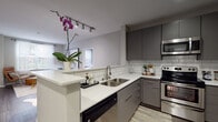 This well-appointed single-floor residence showcases a cohesive modern design with contemporary finishes carried throughout every room. The kitchen is a standout feature, outfitted with flat-front gray cabinetry, white quartz countertops, a classic subway tile backsplash, and a full suite of stainless steel appliances including a range, microwave, refrigerator, and dishwasher. A peninsula with an extended countertop provides additional workspace and connects the kitchen to the living area, which is brightened by a large window dressed with sheer curtains, allowing natural light to fill the space. The living area offers a relaxed yet refined ambiance suited for both everyday comfort and entertaining. The primary bedroom is a serene retreat, featuring plush carpeting, soft neutral tones, and architectural arched alcoves that frame the walls with elegant detail. A second bedroom provides comfortable accommodations with its own cozy, carpet-floored layout. The bathrooms continue the design language of the home, featuring gray vanity cabinetry with white countertops, wood-look flooring, large mirrors, and a soaking tub. The primary bath includes access to a spacious walk-in closet, adding practical storage. Throughout the residence, the interplay of gray tones, clean lines, and modern fixtures creates a polished and cohesive aesthetic.