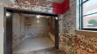 Located at 101–125 E Morehead St in Burlington, NC, this single-floor unit offers a compelling mix of industrial heritage and contemporary design. The space is distinguished by its exposed brick walls, which carry visible layers of history through peeling paint and raw masonry, providing a striking textural contrast to the clean, modern interior finishes found throughout.

The kitchen is a functional centerpiece, featuring dark espresso cabinetry, granite countertops, a peninsula with an integrated double sink, and a full suite of stainless steel appliances including a refrigerator, range, and built-in microwave. Pendant lighting above the peninsula adds warmth and definition to the space. Concrete floors run throughout, lending an industrial aesthetic that ties the historic and modern elements together cohesively.

The unit includes two full bathrooms, each finished with dark vanity cabinetry, granite countertops, and tub-shower combinations. One bathroom features accessibility grab bars. The living and bedroom areas are complemented by ceiling fans, smooth painted walls, and ample room to configure furnishings flexibly. A raw, brick-walled room accessible through a steel-framed opening offers additional utility or creative use potential. Overall, the space delivers a distinctive urban loft character rooted in Burlington's architectural past.