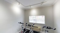 This single-floor fitness space is designed as a dedicated cycling and cardio room. The interior features smooth, light grey walls accented by clean white crown moulding, lending the space a polished, well-maintained appearance. A row of stationary cycling bikes lines the room, providing a focused workout environment suited for spinning sessions or individual cardio training.

Track lighting runs along the ceiling, offering adjustable, targeted illumination throughout the room. A single window fitted with white horizontal blinds allows natural light to filter in, offering a glimpse of surrounding greenery and architecture outside. Beneath the window sits a wall-mounted HVAC unit, ensuring climate control for comfortable year-round use.

The flooring is finished in a dark, low-pile carpet well-suited for exercise equipment placement and easy maintenance. The overall ambiance is clean and functional, with a neutral color palette that keeps the focus on the fitness equipment. The space is efficiently laid out to maximize the number of cycling stations while maintaining an open, uncluttered feel.