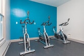 Located at 3900 George Busbee Pkwy NW in Kennesaw, Georgia, this single-floor fitness center offers a well-appointed workout facility designed to support a variety of exercise routines. The main exercise area features bold teal accent walls, rubber flooring, and elevated tray ceilings that lend a sense of height and openness. This space is equipped with multiple treadmills, elliptical machines, a stationary bike, and a multi-function cable strength machine, providing comprehensive cardio and resistance training options. A wall-mounted television adds convenience for those looking to stay entertained during workouts. Large French doors open directly to an outdoor pool area, flooding the space with natural light and offering easy indoor-outdoor access. An adjoining studio room transitions to warm hardwood flooring and provides a flexible open area ideal for stretching, yoga, or functional training. This room includes a punching bag, suspension training equipment, and additional strength accessories mounted along the teal-painted wall. Windows throughout both areas frame views of the surrounding greenery, enhancing the bright and motivating atmosphere. The overall design balances energetic aesthetics with practical functionality, creating a well-rounded fitness environment.