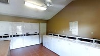 This single-floor communal laundry facility is designed with practicality and efficiency in mind. The room features a row of top-loading washing machines lined along one wall, providing ample capacity for multiple users simultaneously. Complementing these are stacked dryer units positioned along an adjacent wall, maximizing the use of vertical space and overall throughput. The warm, earth-toned walls lend a welcoming feel to what is typically a utilitarian space. Flooring consists of durable tile in a neutral terracotta tone, well-suited to the demands of a high-traffic laundry environment. Overhead, a ceiling fan works in tandem with fluorescent lighting to ensure the room remains bright and well-ventilated during use. Informational signage is posted on the wall, offering washing and drying instructions or guidelines for users. A standard interior door provides access to an adjoining area or utility space. The layout is thoughtfully organized to allow easy movement between machines, making the laundry process straightforward and accessible. Overall, the facility presents a clean, functional, and orderly environment suited for shared residential or commercial use.