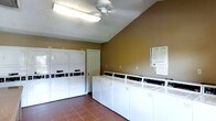This single-floor communal laundry facility is designed with practicality and efficiency in mind. The room features a row of top-loading washing machines lined along one wall, providing ample capacity for multiple users simultaneously. Complementing these are stacked dryer units positioned along an adjacent wall, maximizing the use of vertical space and overall throughput. The warm, earth-toned walls lend a welcoming feel to what is typically a utilitarian space. Flooring consists of durable tile in a neutral terracotta tone, well-suited to the demands of a high-traffic laundry environment. Overhead, a ceiling fan works in tandem with fluorescent lighting to ensure the room remains bright and well-ventilated during use. Informational signage is posted on the wall, offering washing and drying instructions or guidelines for users. A standard interior door provides access to an adjoining area or utility space. The layout is thoughtfully organized to allow easy movement between machines, making the laundry process straightforward and accessible. Overall, the facility presents a clean, functional, and orderly environment suited for shared residential or commercial use.