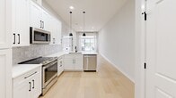 Located at 9 John St in Clinton, CT, this thoughtfully designed one-bedroom, one-bathroom residence offers a clean, modern aesthetic throughout its efficiently utilized single-floor layout. Light-toned hardwood flooring flows seamlessly across the living areas and bedroom, creating a warm, cohesive feel. The kitchen is a standout feature, showcasing white shaker-style cabinetry with matte black hardware, a classic subway tile backsplash, white quartz countertops, and a full suite of stainless steel appliances including a range, built-in microwave, and dishwasher. Pendant lighting above the kitchen and recessed ceiling lights add both functionality and style, while a window at the end of the kitchen invites natural light. The bedroom offers a comfortable footprint with a large window framing outdoor views, providing ample natural light. A closet with double panel doors provides convenient storage directly off the main entry area. The bathroom is finished with large-format neutral floor tiles, a soaking tub and shower combo enclosed by a frameless sliding glass door with matte black hardware, and a white vanity with matching black accents. The overall palette of soft whites and light wood tones gives the space a clean, contemporary ambiance throughout.