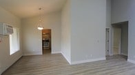 This single-floor space offers a clean, modern aesthetic defined by its airy, open layout and thoughtful design details. The main living area immediately impresses with its soaring vaulted ceilings, which lend a generous sense of height and openness throughout. Smooth, light-toned hardwood flooring runs seamlessly across the space, contributing to a warm yet contemporary feel.

A stylish pendant light fixture serves as a subtle focal point, elegantly bridging the living area to the adjacent kitchen. The kitchen is accessible through a defined entryway and features warm wood cabinetry paired with stainless steel appliances, including a range and oven, creating a functional and visually cohesive cooking environment. A range hood is mounted above the stove, adding both practicality and a polished finish to the culinary space.

The neutral, light grey wall palette throughout enhances the brightness of the interior and provides a versatile backdrop adaptable to a variety of interior styles. White baseboards offer a crisp contrast against the flooring, reinforcing the clean lines of the overall design. The combination of high ceilings, open sightlines, and a cohesive material palette makes this a welcoming and highly functional space.