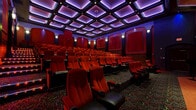 Located at 325 W Adams Blvd in Los Angeles, CA, this dedicated cinema screening room delivers an immersive, theatrically designed entertainment experience across a single, generously sized floor. The auditorium-style layout accommodates multiple rows of plush, red upholstered seats with black leather accents and built-in cup holders, all arranged to face a large stage-front screen concealed behind dramatic floor-length red curtains trimmed with gold fringe. The elevated rear seating rows, accessible via illuminated steps, provide clear sightlines throughout the room.

The coffered ceiling is a standout architectural feature, composed of a grid of recessed panels bathed in vivid purple ambient lighting that casts a rich, cinematic atmosphere across the entire space. Deep red paneled walls adorned with arched moldings and wall-mounted sconces reinforce the classic movie palace aesthetic, while ornate patterned carpeting in rich jewel tones covers the floor, adding to the luxurious ambiance.

Near the entrance, a set of arched wooden doors provides access to the space, accompanied by a podium and stanchion for organized entry. Security and occupancy notices are posted near the entrance, indicating a professionally managed venue. Every design element — from the layered lighting to the theatrical curtain and refined wall treatments — works cohesively to create an intimate yet grand cinematic environment.