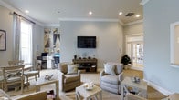 Located at 1600 Belle Point Dr in Mt. Pleasant, South Carolina, this single-floor space offers a well-appointed and welcoming interior designed with both comfort and functionality in mind. The main lounge area is finished with soft, neutral tones — pale gray walls, light tile flooring, and crisp crown molding — creating a calm, coastal-inspired ambiance. A wall-mounted flat-screen television serves as a focal point, complemented by a rustic media console adorned with tasteful decorative accents. Comfortable upholstered armchairs are arranged around a pair of low coffee tables, providing an inviting setting for relaxation or casual gatherings. Recessed lighting illuminates the space evenly, enhancing the airy feel of the room. Adjacent to the lounge, a dedicated workstation area is equipped with a desk, computer, and ergonomic seating, making the space equally suited for productivity. Large French doors allow natural light to filter in and offer a view to the exterior, further brightening the interior. Decorative artwork and potted greenery add warmth and personality throughout, rounding out the polished yet approachable character of this thoughtfully designed space.