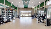 This single-level fitness center is designed to accommodate a wide range of workout styles within a spacious, open-concept layout. The space is divided into distinct functional zones, including a cardio area outfitted with multiple treadmills and rowing machines, a strength training section featuring cable machines and a multi-rack weight station, and an open floor area suitable for functional or group training.

Architecturally, the facility stands out with soaring ceilings accented by a tray detail finished in teal-green, complemented by a large ceiling fan that promotes air circulation. Bold green wall panels and a striking chevron-patterned accent wall add visual energy to the interior. Full-length mirrors line portions of the walls, enhancing the sense of space and providing visual feedback during workouts.

Natural light is a defining feature of this fitness center. A full-width glass panel garage door on one wall floods the training floor with daylight and offers views of an outdoor patio area. Additional large windows along the perimeter further brighten the space. The durable, light-toned flooring with dark rubber inlay strips provides both practicality and a clean aesthetic throughout the entire facility.