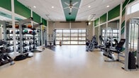 This single-level fitness center is designed to accommodate a wide range of workout styles within a spacious, open-concept layout. The space is divided into distinct functional zones, including a cardio area outfitted with multiple treadmills and rowing machines, a strength training section featuring cable machines and a multi-rack weight station, and an open floor area suitable for functional or group training.

Architecturally, the facility stands out with soaring ceilings accented by a tray detail finished in teal-green, complemented by a large ceiling fan that promotes air circulation. Bold green wall panels and a striking chevron-patterned accent wall add visual energy to the interior. Full-length mirrors line portions of the walls, enhancing the sense of space and providing visual feedback during workouts.

Natural light is a defining feature of this fitness center. A full-width glass panel garage door on one wall floods the training floor with daylight and offers views of an outdoor patio area. Additional large windows along the perimeter further brighten the space. The durable, light-toned flooring with dark rubber inlay strips provides both practicality and a clean aesthetic throughout the entire facility.