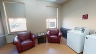 This single-floor laundry facility is designed to combine practicality with a degree of comfort, making it a well-rounded communal space. The room is finished with neutral beige walls and durable light-toned flooring, creating a clean and welcoming atmosphere throughout.

The laundry area is equipped with multiple top-loading washing machines, clearly labeled and arranged neatly along one wall. Caution signage is posted above each unit, reflecting an attention to user safety. A heating unit is also present, ensuring the space remains comfortable year-round.

A dedicated seating area occupies one portion of the room, furnished with two plush burgundy leather armchairs arranged around a small dark wood side table stocked with reading materials. This thoughtful addition provides a relaxed spot for occupants to wait during laundry cycles.

Natural light fills the space through several windows fitted with roller shades, offering views of the surrounding streetscape, including glimpses of neighboring brick buildings and street-level details. The elevated window placement contributes to the room's open, airy feel without compromising privacy.

Overall, this laundry facility strikes a balance between utility and comfort, with its tidy layout, ample equipment, and inviting seating area making it a practical and pleasant shared space.