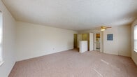This single-floor residence offers a practical and comfortable layout designed for everyday living. The expansive living area is finished with soft wall-to-wall carpeting, neutral cream-toned walls, and a textured ceiling, creating a bright and open atmosphere. A ceiling fan provides overhead circulation, while multiple windows along one wall allow generous natural light to fill the space. A half-wall partition separates the living area from the kitchen, maintaining an open connection between the two spaces. The kitchen is well-equipped with warm honey-toned wood cabinetry extending to the upper walls, offering ample storage. Light-toned laminate countertops, a stainless steel sink, a gas range, and a full-size refrigerator complete the functional cooking space. Tile flooring runs throughout the kitchen for easy maintenance. The bedroom features plush carpeting, a ceiling light fixture, a built-in closet, and a wall-mounted HVAC unit beneath dual windows that look out to greenery outside. The full bathroom includes tile flooring, tile wainscoting, a combined tub and shower, a toilet, and a vanity with a wood cabinet base, all brightened by a window with outdoor views.