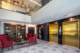 Located at 839 Sunrise Hwy in Lynbrook, New York, this elegantly designed lobby presents a refined first impression through its thoughtful architectural details and upscale finishes. The space is anchored by expansive marble tile flooring with bold veining that flows seamlessly throughout, lending a sense of grandeur and cohesion to the entire level.

Upon entry, floor-to-ceiling black-framed glass doors flood the foyer with natural light and offer views to the exterior. A concierge desk positioned nearby provides a functional yet stylish welcome point. Two distinct seating areas define the lobby's character: one features a cluster of bold crimson wingback chairs arranged around a sleek black coffee table, set against a built-in walnut shelving unit displaying curated décor and a striking piece of wall art; the other showcases a generous caramel leather sectional sofa paired with a gold-accented chandelier and a wall-mounted display screen, all framed by large windows overlooking the outdoors.

A dramatic elevator bank serves as a focal point, with dual gold-finished elevator doors set against a dark, richly veined marble accent wall and decorative wave-patterned inlays. Overhead, a sculptural gold globe chandelier adds a luxurious finishing touch. The overall ambiance is modern and sophisticated, balancing bold design choices with welcoming, functional spaces.