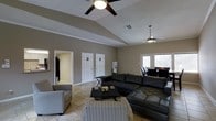 This single-floor space is designed with an open and inviting layout that maximizes comfort and functionality. The main living area is a standout, boasting soaring vaulted ceilings that lend a sense of spaciousness and airiness to the room. Two ceiling fans are installed — one centrally positioned over the main seating area and another toward the rear — ensuring excellent air circulation throughout.

The living area is appointed with a large sectional leather sofa, an accent armchair with a patterned fabric, and a matching patterned ottoman, all arranged around a sleek black coffee table. The neutral taupe wall color complements the light beige tile flooring, creating a cohesive and understated aesthetic.

Natural light filters in through a set of double windows along one wall, brightening the space without overwhelming it. A glimpse into the adjacent dining area reveals additional seating and table space, suggesting a smooth transition between living and dining zones.

Several white interior doors along the back wall lead to additional rooms or utility areas, maintaining a clean and organized appearance. The overall ambiance is warm and practical, with architectural details like vaulted ceilings and thoughtful furnishing choices combining to create a welcoming, well-appointed environment.