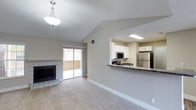 This single-floor residence showcases a thoughtfully designed open-concept layout that seamlessly connects the living area and kitchen. The living room draws immediate attention with its vaulted ceiling, which adds a sense of spaciousness and airiness to the space. A pendant light fixture provides warm, centralized illumination, while the textured ceiling and neutral-toned walls contribute to a clean, inviting atmosphere.

A tile-surround fireplace anchors one wall of the living area, serving as a functional focal point that adds both character and comfort. Plush wall-to-wall carpeting runs throughout the living space, enhancing the room's warmth. A sliding glass door leads directly to a private balcony, allowing for natural light to fill the interior and offering easy outdoor access.

The kitchen is positioned adjacent to the living area, separated by a raised breakfast bar with a dark granite-style countertop — ideal for casual dining or entertaining. White cabinetry lines the walls, complemented by stainless steel appliances including a refrigerator, microwave, and range. Recessed overhead lighting keeps the kitchen bright and functional. Together, these elements create a cohesive, comfortable living environment that balances practicality with understated style.