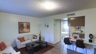 This single-floor unit offers a well-organized layout that maximizes its available space. The living area serves as the central hub, featuring wood-style flooring, a textured ceiling, and neutral white walls that create a clean, bright atmosphere. A comfortable sofa with decorative cushions anchors the space, complemented by a dark wood coffee table and a patterned area rug that adds warmth underfoot. Subtle wall décor, including a framed artwork piece and decorative candle sconces, contribute to a modern yet cozy aesthetic.

Adjacent to the living area is a compact dining space, furnished with a sleek dark table and contemporary chairs, making efficient use of the available floor plan. A kitchen area is partially visible, featuring dark wood cabinetry and standard appliances.

Through an open doorway, the bedroom is visible, benefiting from natural light supplied by a window. The room accommodates a full-sized bed and maintains the same neutral color palette seen throughout the unit.

Overall, the space presents a cohesive and functional layout with consistent flooring, a neutral color scheme, and practical furnishings that make efficient use of every area.