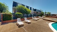 This outdoor amenity area at 2436 Beach Blvd in Biloxi, Mississippi, centers around a generously sized kidney-shaped swimming pool with clear turquoise water, surrounded by an expansive concrete and aggregate sun deck. The pool area is well-appointed with numerous white lounge chairs positioned throughout the deck, offering ample space for sunbathing and relaxation. A dedicated seating area with a round table and chairs provides a comfortable spot for poolside gathering.

The space is framed by a classic brick building featuring distinctive arched openings, a slate-toned roof, and a chimney, which serves as a pool house or utility structure. A wrought iron fence with brick pillars encloses the area, providing both security and a polished aesthetic. The surrounding multi-story brick buildings with dark mansard-style rooflines and private balconies add architectural character to the setting.

Mature trees and manicured shrubs border the perimeter, lending greenery and natural shade. The entire outdoor space is finished with textured aggregate concrete paving, and tall light poles ensure evening usability. The overall ambiance is inviting and resort-like, well-suited to the Gulf Coast setting.