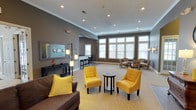 This single-floor space is designed as an open, welcoming lounge area with a thoughtfully curated interior that balances comfort and style. The room is finished with neutral gray walls and a textured ceiling punctuated by recessed lighting, which works alongside wall-mounted sconces to create a warm, well-lit atmosphere throughout the day and evening.

The main seating area is anchored by a pair of bold yellow tufted accent chairs arranged around a small round side table, complemented by a brown upholstered sofa with a matching yellow throw pillow — creating a vibrant yet cohesive color palette. A large decorative mirror and a console table along one wall add a refined touch, while a tall multi-pendant floor lamp provides additional ambient lighting.

Toward the rear of the space, a secondary seating arrangement features patterned upholstered chairs positioned near a row of large, multi-pane windows that flood the area with natural light and offer views of the surrounding landscape. A bar-height counter with stools is visible in the background, suggesting a refreshment or café-style nook. The open layout and generous proportions give the space an airy, communal feel suitable for gathering and relaxation.