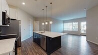 Located at 1145 Chambers Rd in Columbus, Ohio, this single-floor residence showcases a contemporary design with thoughtfully selected finishes throughout. The open-concept kitchen serves as the centerpiece of the living space, anchored by a large island with a navy blue base, white quartz countertops, and an integrated sink with a brushed gold faucet. White shaker-style cabinetry lines the walls above a classic white subway tile backsplash, complemented by a full suite of stainless steel appliances including a range, microwave, and refrigerator. Globe pendant lights with brass accents hang above the island, adding a warm, modern touch. Dark hardwood-style flooring extends throughout the kitchen and main living areas, creating a cohesive and grounded aesthetic. The living space opens to a windowed door providing natural light and outdoor access. Bedrooms feature plush carpeting and additional exterior-facing doors. The bathrooms are well-appointed: one includes a glass-enclosed walk-in shower with white subway tile and a navy vanity, while another offers a full tub-shower combination with oil-rubbed bronze fixtures. Neutral wall tones throughout contribute to a clean, move-in-ready atmosphere.