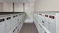 This postal center is a single-floor facility designed to efficiently serve residents' mail and package needs. The space is organized into distinct functional areas, beginning with a welcoming entry vestibule featuring large windows that allow natural light to filter in and offer views of the surrounding neighborhood. The entry area includes a waste receptacle and a wall-mounted fire extinguisher, reflecting the facility's attention to safety and cleanliness.

The primary mail room houses extensive rows of individually numbered, gray metal mailboxes arranged in neat parallel columns along both walls, creating a narrow, organized corridor. Each box is clearly labeled with a numbered placard for easy identification. Overhead fluorescent lighting ensures consistent illumination throughout this corridor.

A separate alcove area accommodates oversized parcel lockers in a tan/beige finish, providing secure package retrieval for larger deliveries. These lockers are tiered, offering multiple size options. The flooring throughout is durable, low-pile carpet suitable for regular foot traffic. Signage throughout the facility communicates property rules, no-smoking policies, and surveillance notifications, contributing to a secure, well-managed environment.