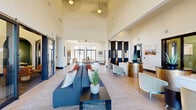 This expansive single-floor amenity facility is a masterclass in contemporary design, thoughtfully curated to offer a variety of social and recreational experiences. The grand entrance lobby immediately impresses with soaring ceilings, statement pendant light fixtures, and light herringbone hardwood flooring that flows throughout the main areas. A welcoming reception desk, flanked by elegant brushed-gold accent pedestals and lush tropical plants, sets the tone for the refined aesthetic found throughout.

The open-plan main lounge accommodates multiple seating arrangements, bathed in natural light from floor-to-ceiling windows and glass doors that open to outdoor views. An adjoining kitchen and dining area features exposed beam ceilings, warm wood cabinetry, a kitchen island with bar seating, and generous dining tables — ideal for community gatherings.

A dedicated games room offers a full-size billiards table, plush lounge seating, and a large wall-mounted display, all beneath dramatic cone pendant lighting. An adjacent café-style lounge presents intimate round tables, leather chairs, a slatted wood accent wall with a mounted screen, and a charming arched alcove for private seating.

Completing the amenity offering is a sleek screening room, finished in deep charcoal tones with a large projection screen, tiered seating, and warm ambient wall sconces — delivering a true cinematic experience.