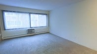 This single-floor space presents a clean, functional living area characterized by its simple, understated design. The room features wall-to-wall carpeting in a neutral tone, providing a soft and comfortable foundation throughout. A large window dominates one wall, fitted with vertical blinds that allow for adjustable natural light control, while offering a view of the surrounding exterior. Beneath the window, a wall-mounted air conditioning unit ensures climate comfort, accompanied by a baseboard heating system that runs along the lower perimeter of the wall — a practical combination for year-round temperature regulation. The walls are finished in a bright, neutral white, contributing to an open and airy atmosphere that makes the space feel clean and versatile. The overall layout is open and unfurnished, allowing for flexible arrangement of furniture and décor to suit a variety of needs. The space is straightforward in its design, prioritizing functionality while maintaining a bright and livable ambiance suitable for everyday use.