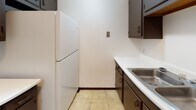 This single-floor unit at 2824 Airport Hwy in Toledo, Ohio offers a straightforward and functional layout designed for comfortable everyday living. The spacious living area features neutral-toned carpet, white walls, and a textured ceiling, creating a clean and open atmosphere. A wall-mounted air conditioning unit and baseboard heating provide year-round climate control, while a window with blinds allows for natural light and ventilation. The kitchen is efficiently arranged with dark wood cabinetry, white countertops, a stainless steel sink, and tile flooring. A full-size refrigerator is included, and the galley-style layout makes practical use of the available space. The bedroom is generously sized with soft carpeting, white walls, and a large sliding closet offering substantial storage. An additional closet with paneled doors is also present in the hallway area, providing further organizational space. The bathroom is neatly appointed with light wood-look flooring, a dark vanity with a light countertop, a well-lit mirror, and a full bathtub with a shower rod. A linen closet adjacent to the bathtub adds convenient storage. Overall, the unit presents a clean, practical living environment suited for those seeking a functional and move-in ready space in Toledo.