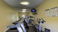 This fitness center is a dedicated, single-floor workout facility designed to meet a range of exercise needs. The space is outfitted with an array of cardio equipment, including treadmills, elliptical trainers, and stationary bikes, arranged to allow comfortable movement between machines. A wall-mounted flat-screen television is positioned for easy viewing during workouts, adding an element of entertainment to exercise sessions. Mirrored walls line one side of the room, creating a sense of added depth while allowing users to monitor their form. The ceiling features flush-mounted circular light fixtures that provide consistent, even illumination throughout the space. The walls are painted in a soft yellow tone, contributing to a bright and energizing atmosphere. A textured ceiling and tile flooring complete the practical, functional aesthetic of the room. Additional strength and multi-use equipment can be seen toward the back of the space, making this a comprehensive facility suited for both cardio and resistance training. The overall layout is efficient and well-organized, making the most of the available space for a focused fitness environment.