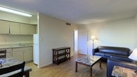 This single-floor space features a straightforward, functional layout designed for comfortable everyday living. The living area is finished with light hardwood-style flooring that extends throughout, providing a warm and cohesive look. Neutral beige walls and a textured ceiling contribute to a clean, understated aesthetic. The living room accommodates a leather sofa, a marble-top coffee table, and a shelving unit, with a floor lamp offering ambient lighting. HVAC vents are positioned on both the wall and ceiling, ensuring climate control throughout the space. A doorway leads from the living area to an additional room, likely a bedroom, enhancing the sense of flow between spaces. To the left, a partial view into the kitchen reveals a white refrigerator and cabinetry, suggesting a compact but practical cooking area. The overall ambiance is modest and utilitarian, with a simple design that prioritizes function. Natural light appears limited based on the visible interior, with artificial lighting supplementing the space. The layout makes efficient use of the available footprint, offering a manageable and easy-to-maintain living environment.