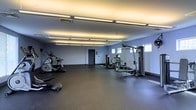 Located at 205 S Front St in the heart of Columbus, Ohio, this fitness center offers a dedicated space for health and wellness. The single-floor facility is designed with functionality and comfort in mind, featuring smooth-painted light gray walls that create a clean, focused atmosphere throughout.

The room is generously sized, providing ample open floor space for movement and exercise. Durable dark rubber flooring covers the entire area, offering both safety and practicality underfoot. Overhead, rows of long fluorescent strip lights are mounted flush against the ceiling, delivering consistent, bright illumination across the entire space.

The facility is equipped with a well-rounded selection of exercise machines, including an elliptical trainer and a multi-station strength training machine, catering to both cardio and resistance training needs. Additional equipment is spread throughout the open floor, allowing for a versatile workout experience.

Natural light filters in through a set of windows along one wall, complementing the overhead lighting and contributing to an open, energizing environment. A secure access door provides a private entrance to the space. Overall, this fitness center strikes a balance between practicality and comfort, making it a well-appointed destination for everyday fitness routines.