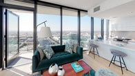 This contemporary high-rise residence is designed with a clean, modern aesthetic throughout its single-floor layout. The living area serves as the centerpiece of the space, anchored by expansive floor-to-ceiling windows on two sides that frame sweeping panoramic city views, flooding the room with abundant natural light. Light hardwood flooring runs throughout, complementing the crisp white walls and creating a warm, airy atmosphere.

The kitchen is outfitted with high-gloss white cabinetry extending to the ceiling, sleek white quartz countertops, a stainless steel undermount sink, and stainless steel appliances, including a gas range and a full-size refrigerator. The minimalist design maximizes both storage and workspace efficiency.

The bedrooms offer a tranquil retreat. The primary bedroom features dramatic floor-to-ceiling windows with commanding city views, while a secondary bedroom includes a dedicated walk-in closet with built-in shelving. Both rooms continue the light hardwood flooring and neutral palette.

Two well-designed bathrooms complete the space. The primary bathroom features a glass-enclosed shower adorned with hexagonal mosaic tile, a separate soaking tub set into marble-style surround, and polished concrete flooring. The secondary bathroom offers a tub-shower combination with large-format marble tile, a floating white vanity, and an illuminated mirror, delivering a refined, hotel-inspired finish throughout.