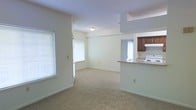 This single-floor unit is designed around an open-concept layout that seamlessly connects the main living area to the kitchen. The living space is generously proportioned, finished with wall-to-wall carpet in a neutral beige tone that contributes to a warm, understated atmosphere. Cream-colored walls and a flush ceiling light fixture keep the space feeling clean and bright.

Natural light enters through two windows fitted with horizontal blinds — one positioned along the side wall and another near what appears to be an entry or exterior door — providing adequate illumination throughout the day. A doorway with white trim leads further into the unit, suggesting additional rooms beyond the main living area.

The kitchen is partially open to the living space via a raised counter-height bar, creating a functional divide while maintaining visual connectivity. Dark wood cabinetry lines the upper portion of the kitchen, complemented by a white range and range hood. The layout is compact yet practical, offering essential cooking and storage amenities.

Overall, the space presents a straightforward, move-in-ready design with a cohesive neutral palette suitable for a variety of furnishing styles.