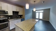 This single-floor space showcases a thoughtfully designed open-concept layout that seamlessly connects the kitchen and living area. The kitchen is a standout feature, anchored by a generous granite-topped island with warm, speckled tones that serves as both a workspace and a gathering point. White cabinetry lines the walls, providing ample storage while contributing to a clean, bright aesthetic. A textured dark tile backsplash adds visual contrast behind the sink area, and stainless steel appliances, including a dishwasher, complete the functional cooking space. Two cylindrical pendant lights hang above the island, casting a warm, focused glow that enhances the kitchen's inviting atmosphere. The living area flows naturally from the kitchen, featuring soft blue-gray walls that create a calm, neutral backdrop. Dark hardwood flooring runs throughout, grounding the space with a rich, cohesive tone. At the far end of the living area, a set of French doors fitted with horizontal blinds allows an abundance of natural light to filter in while offering a connection to the outdoors. The overall ambiance is modern and clean, balancing practical design with an open, comfortable feel.