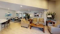 This single-story amenity space is thoughtfully designed to provide a welcoming and functional environment for socializing and relaxation. The interior is finished with light wood-look flooring throughout, recessed lighting, and neutral gray-toned walls that create a clean, contemporary aesthetic.

The lounge area serves as the centerpiece of the space, featuring patterned upholstered armchairs arranged around a solid wood coffee table. Framed artwork and a lush fiddle-leaf fig plant add warmth and visual interest. Large floor-length mirrors with dark frames are strategically placed to enhance the sense of depth and openness throughout the room.

Adjacent to the lounge, a dedicated dining area accommodates multiple white-topped square tables paired with dark gray chairs, suitable for group gatherings. Colorful butterfly artwork decorates the walls, contributing a lively touch to the setting.

The kitchenette features dark laminate cabinetry, a light quartz countertop, a stainless steel refrigerator with ice dispenser, microwave, single-serve coffee maker, and a sink — offering convenient refreshment amenities. Vaulted ceilings above this area provide an airy, open feel.

A separate game nook near the entrance includes an arcade table and bright orange accent chairs. A glass door opens directly to an outdoor patio, filling the space with natural light.