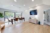 This well-appointed single-floor space showcases a thoughtfully designed open-concept layout that seamlessly connects its key living areas. Consistent light wood luxury vinyl plank flooring flows throughout, lending warmth and visual continuity to every room.

The expansive main living and dining area is anchored by vaulted ceilings and recessed lighting, creating an airy, open atmosphere. The dining space is highlighted by a stylish gold globe pendant chandelier suspended above a large wooden dining table surrounded by upholstered chairs, all positioned in front of oversized black-framed picture windows that frame lush outdoor greenery and fill the room with natural light. A wall-mounted flat-screen TV and a neutral dresser-style credenza define the living zone.

A comfortable sitting room or reception area features large windows dressed with linen drapes and gold curtain rods, flooding the space with soft, diffused light. Cream upholstered accent chairs and a gold side table contribute to the refined, modern aesthetic.

Additional rooms include a dedicated fitness room accessible through a glass-paned French door, and an office or workspace visible through a classic sliding barn door, complete with built-in shelving. The textured ceilings and soft gray-white wall palette throughout the space provide a clean, cohesive backdrop that complements the modern furnishings and gold accent fixtures.