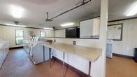This single-floor communal laundry facility is designed to accommodate high-volume use, offering a practical and functional layout. The room is generously sized, housing multiple rows of both top-loading and front-loading washing machines alongside a full complement of dryers, providing ample capacity for shared use. Fluorescent lighting fixtures mounted to the textured ceiling illuminate the space evenly, while ceiling fans aid in air circulation throughout. The flooring throughout is a durable terracotta-toned material suited for heavy foot traffic.

A long folding counter supported by metal legs runs along one side of the room, offering a dedicated surface for sorting and folding laundry. Adjacent to this area, a pass-through window opens to a secondary section of the room where additional appliances, including a large upright refrigerator and a smaller white unit, are stored. White vertical panel wainscoting lines the lower portions of the walls, accented by dark wood trim. Small windows allow modest natural light to enter, and a bulletin board is mounted near one of the entry doors. A standard exterior door with a windowed panel provides access to the outside, completing this straightforward, utilitarian space.