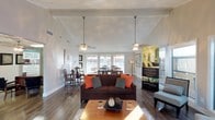 This single-story space showcases an open-concept layout that seamlessly connects the living and dining areas, creating a cohesive and functional environment. The living area is anchored by a dark brown sofa accented with vibrant orange and teal throw pillows, paired with a warm wood coffee table that adds a natural touch to the space. Overhead, vaulted ceilings with elegant tray detailing add a sense of height and architectural interest, while two pendant light fixtures with ceiling fan elements provide both illumination and airflow.

The hardwood flooring runs continuously throughout the space, unifying the living and dining zones. The dining area, positioned toward the rear of the space, features a classic wood table with traditional-style chairs, bathed in natural light streaming through large windows and sliding glass doors. These generous openings not only flood the interior with daylight but also offer views to the outdoor area beyond.

The neutral wall tones and clean architectural lines create a bright, airy ambiance throughout, while the carefully chosen furnishings and décor add warmth and character. The overall design balances comfort and style in a well-proportioned, light-filled setting.