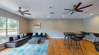 This single-floor recreation room in Jackson, Tennessee, offers a well-organized and inviting communal space designed for relaxation and social interaction. The room features warm-toned hardwood flooring throughout, complemented by neutral gray walls and recessed lighting, creating a bright and welcoming atmosphere. Two ceiling fans help maintain comfortable airflow across the generously sized open floor plan.

The lounge area is anchored by a large dark sectional sofa paired with a woven ottoman coffee table, set atop a vibrant blue area rug. Large multi-pane windows flood this seating zone with natural light and provide views of the surrounding greenery, enhancing the room's open, airy feel. A wall-mounted television is positioned nearby, adding an entertainment element to the space.

The dining and workspace area features a woven high-top table with matching bar-height chairs, alongside a long built-in counter with bar seating — ideal for casual dining or individual work. Framed artwork adds decorative character throughout.

A set of French doors with glass panels leads to the exterior, further connecting the interior to the outdoors. The overall design balances functionality with comfort, making this recreation room a practical and pleasant gathering space.