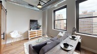 This thoughtfully designed urban unit blends industrial and contemporary aesthetics across a single open floor plan. The living area makes a strong impression with exposed silver ductwork overhead, warm hardwood flooring underfoot, and large black-framed windows that frame sweeping city skyline views and flood the space with natural light. A combined living and dining area provides a comfortable, versatile layout anchored by a plush sectional sofa and a rustic wood dining table.

The kitchen is well-appointed with rich dark wood cabinetry, light quartz countertops, a classic white subway tile backsplash, and a full suite of stainless steel appliances including a professional-grade gas range and built-in microwave. Track lighting illuminates the workspace efficiently.

The bedroom offers a serene retreat, featuring a distinctive herringbone wood headboard and access to the living area through a sliding reclaimed wood barn door. A matching barn door connects to the ensuite bathroom, which is equipped with accessibility features including grab bars, a spacious vanity with dark countertop, white cabinetry, and neutral tile flooring. The overall palette of warm grays, natural wood tones, and matte black accents creates a cohesive, modern aesthetic throughout.