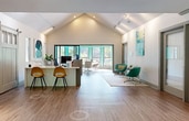 This contemporary 1,420 square foot office space in Alpharetta, Georgia, showcases a harmonious blend of functionality and style. The single-story layout is characterized by an open floor plan that maximizes space utilization. A striking turquoise accent wall serves as a focal point, injecting vibrancy into the predominantly white and wood-toned interior.

The kitchen area boasts modern amenities, including stainless steel appliances, white cabinetry, and a spacious island with bar seating. Pendant lighting with unique glass shades adds a touch of elegance. Adjacent to the kitchen, a dining area with a round table provides a communal space for meals or informal meetings.

A dedicated workstation area features a long white counter with bar-height seating, ideal for collaborative work or individual tasks. The space is illuminated by recessed lighting and large windows that offer views of the surrounding greenery, ensuring ample natural light throughout the day.

The lounge areas are thoughtfully designed with comfortable seating options, including modern armchairs in teal and a neutral sofa. These spaces are perfect for casual meetings or moments of relaxation. Strategic placement of artwork and decorative elements enhances the overall aesthetic appeal.

The office's high ceilings with exposed beams contribute to a sense of spaciousness, while the wood-look flooring adds warmth and durability. The reception area features a sleek white counter and sliding barn doors, blending rustic charm with modern design.

Overall, this office space offers a versatile and inviting environment that caters to various work styles and promotes productivity in a visually appealing setting.