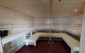 This 150-square-foot sauna room is a single-floor wellness space designed for relaxation and heat therapy. The interior is entirely clad in light, natural pine wood — covering the walls, ceiling, and slatted bench surfaces — creating a warm, cohesive Scandinavian aesthetic. Multi-tiered wooden benches line three walls in an L-shaped and wrap-around configuration, providing ample seating and reclining space for multiple users at varying heat levels. The lower floor area features a deep red interlocking tile surface, offering a practical and slip-resistant contrast to the pale wood above. A sauna heater unit with a stone bed is positioned near the entrance, serving as the heat source for the room. Recessed ceiling spotlights provide subtle, even illumination throughout the space. A glass-paneled wooden door connects the sauna directly to an adjacent fitness center, visible through the glazing, allowing for convenient transitions between workout and recovery. The overall ambiance is clean, minimal, and purpose-built, emphasizing function and comfort in equal measure.