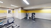 This fitness center is a dedicated, single-floor exercise facility designed to support a range of workout routines. The space is defined by a distinctive interior design featuring gray walls accented by a bold yellow horizontal stripe that runs throughout the room, creating an energetic and motivating atmosphere. The durable rubber flooring provides a safe, cushioned surface suitable for all types of physical activity.

The facility is generously equipped with a variety of cardio machines, including treadmills, elliptical trainers, and stationary bikes — featuring brands such as Spirit and Precor. A multi-function cable machine rounds out the strength-training options, catering to both beginner and experienced gym-goers. A seating area with shelving near the exit door offers a convenient spot to rest or store personal belongings.

Mirrored walls along one side of the room enhance the sense of openness and allow users to monitor their form during workouts. Natural light filters in through several windows along the walls, supplemented by flush-mounted fluorescent ceiling fixtures that ensure the space remains bright at all hours. Wall-mounted air conditioning units provide climate control throughout. The layout is open and thoughtfully organized, allowing for comfortable movement between equipment.