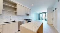 Located on the 17th floor in the heart of downtown San Diego (92101), this modern high-rise residence offers a thoughtfully designed single-level layout with clean contemporary aesthetics throughout. Light-toned hardwood flooring flows seamlessly across the open-concept living and kitchen areas, creating a cohesive and airy atmosphere.

The kitchen is a standout feature, equipped with a large waterfall-edge quartz island, flat-front cabinetry in warm wood and matte finishes, white subway tile backsplash, and a full suite of stainless steel appliances including a gas range, refrigerator, built-in microwave, and dishwasher. Ample upper cabinetry with open shelving provides generous storage.

The living area opens directly to a private balcony through floor-to-ceiling black-framed glass doors, filling the space with natural light and offering sweeping views of the San Diego skyline and surrounding cityscape. Exposed concrete structural columns add an industrial accent against the crisp white walls.

Bedrooms feature the same light hardwood flooring, large windows with city views, built-in shelving, and barn-style sliding doors. The primary bathroom is finished with large-format white tile flooring, a double vanity with quartz countertops, backlit mirrors, wood cabinetry with brushed gold hardware, and a soaking tub alongside a tiled shower surround.